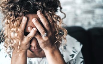 A woman with curly hair is sitting, covering her face with both hands. She is wearing a white patterned blouse and has rings on her fingers. The background is a blurred wall, suggesting an indoor setting.