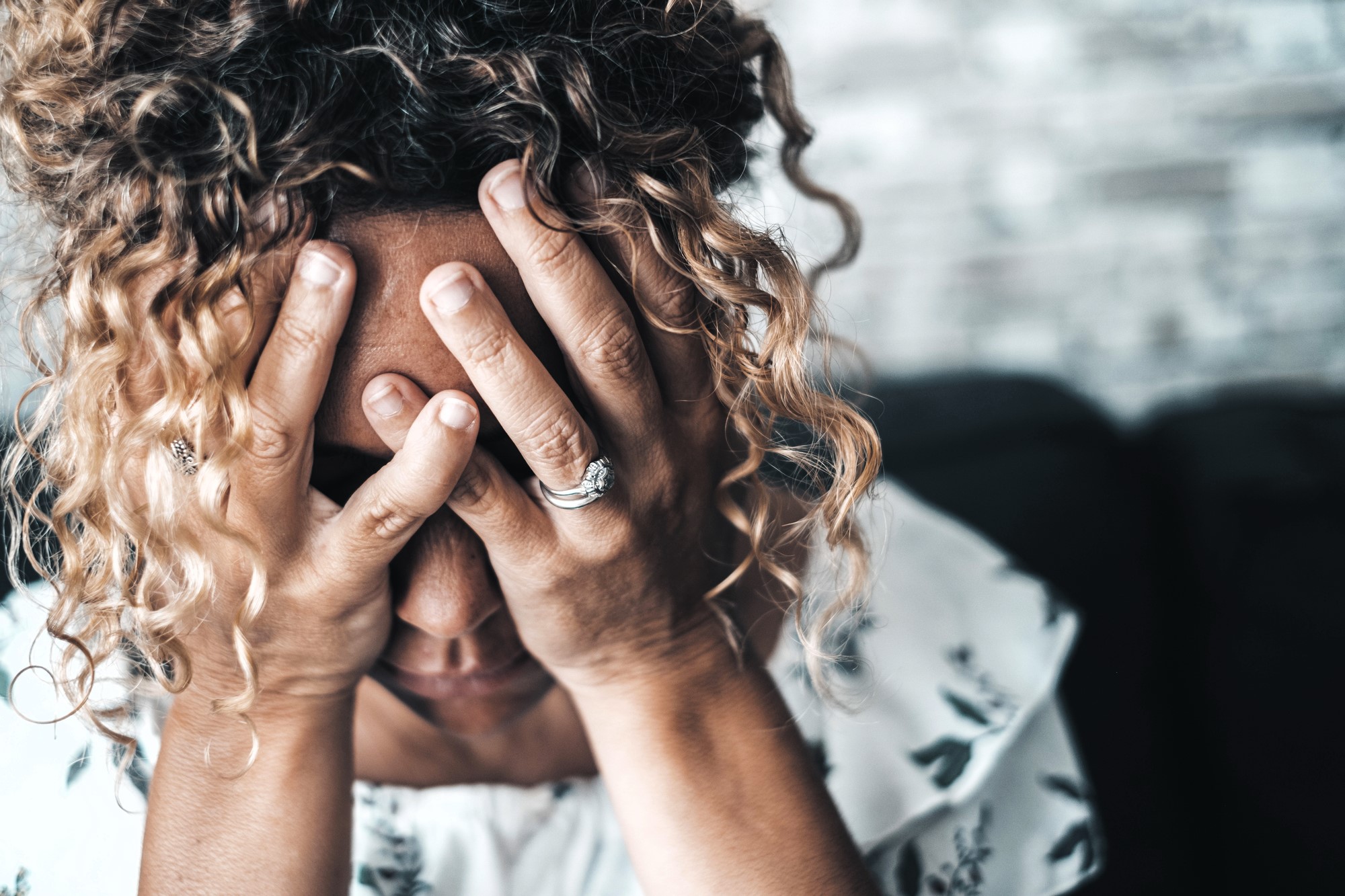 A woman with curly hair is sitting, covering her face with both hands. She is wearing a white patterned blouse and has rings on her fingers. The background is a blurred wall, suggesting an indoor setting.