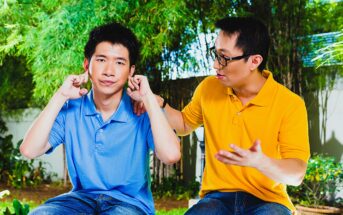 Two men sit outdoors; one in a blue shirt covering his ears with his fingers and looking indifferent. The other in a yellow shirt is gesturing and appears to be talking to him. Lush greenery is visible in the background.