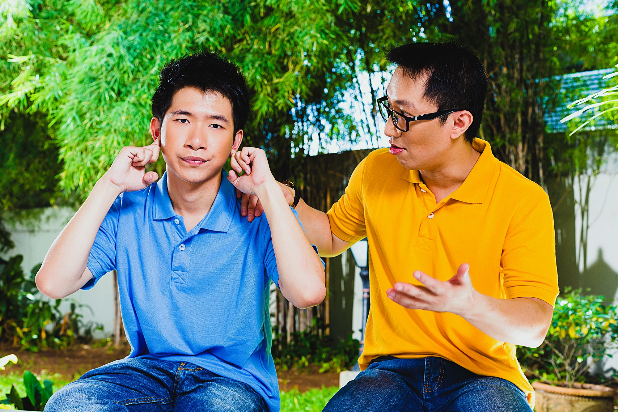 Two men sit outdoors; one in a blue shirt covering his ears with his fingers and looking indifferent. The other in a yellow shirt is gesturing and appears to be talking to him. Lush greenery is visible in the background.