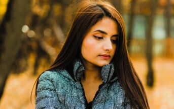 A woman with long hair and winged eyeliner stands outdoors, wearing a patterned jacket. She looks down thoughtfully, with a blurred background of trees in warm autumn colors.