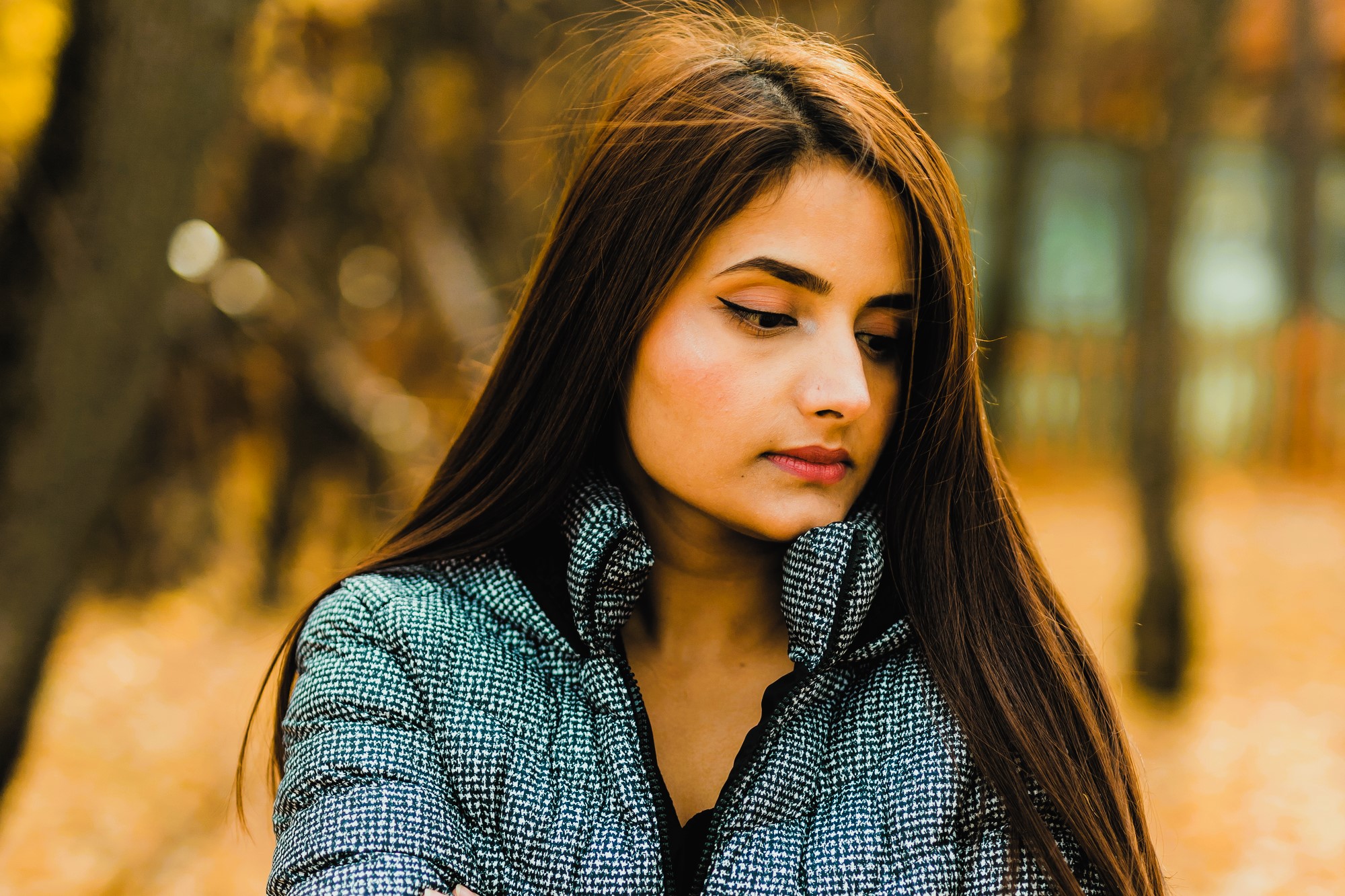 A woman with long hair and winged eyeliner stands outdoors, wearing a patterned jacket. She looks down thoughtfully, with a blurred background of trees in warm autumn colors.