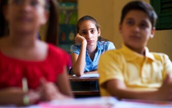 A girl in a classroom, wearing a blue shirt, rests her chin on her hand and looks bored as two classmates in the foreground, wearing a red and a yellow shirt, are blurred, suggesting they might be talking or listening.