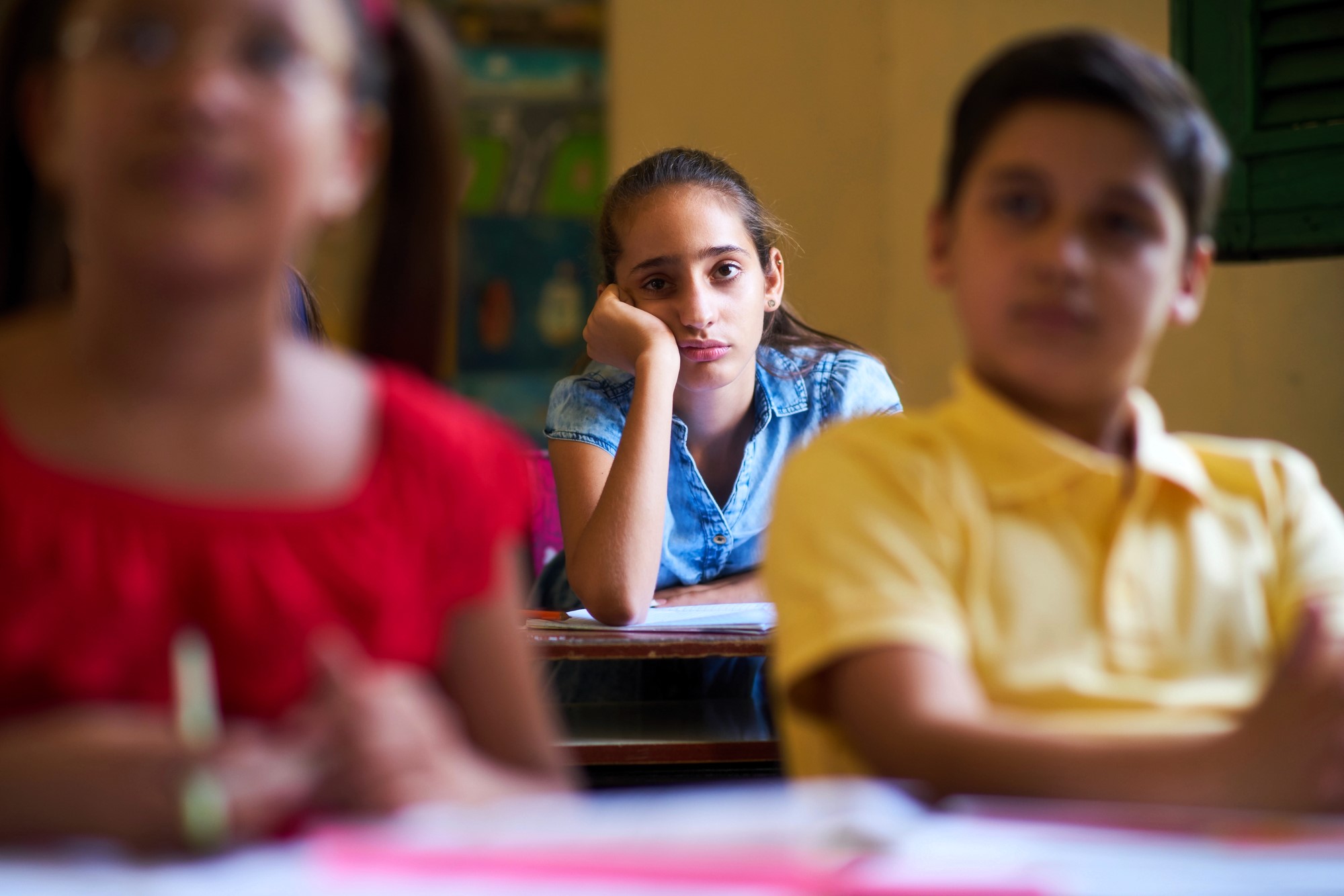 A girl in a classroom, wearing a blue shirt, rests her chin on her hand and looks bored as two classmates in the foreground, wearing a red and a yellow shirt, are blurred, suggesting they might be talking or listening.