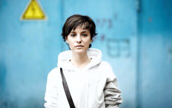 A person with short, dark hair wearing a white hoodie and a black strap across their shoulder stands in front of a blue wall with a yellow warning sign. The background is slightly out of focus.