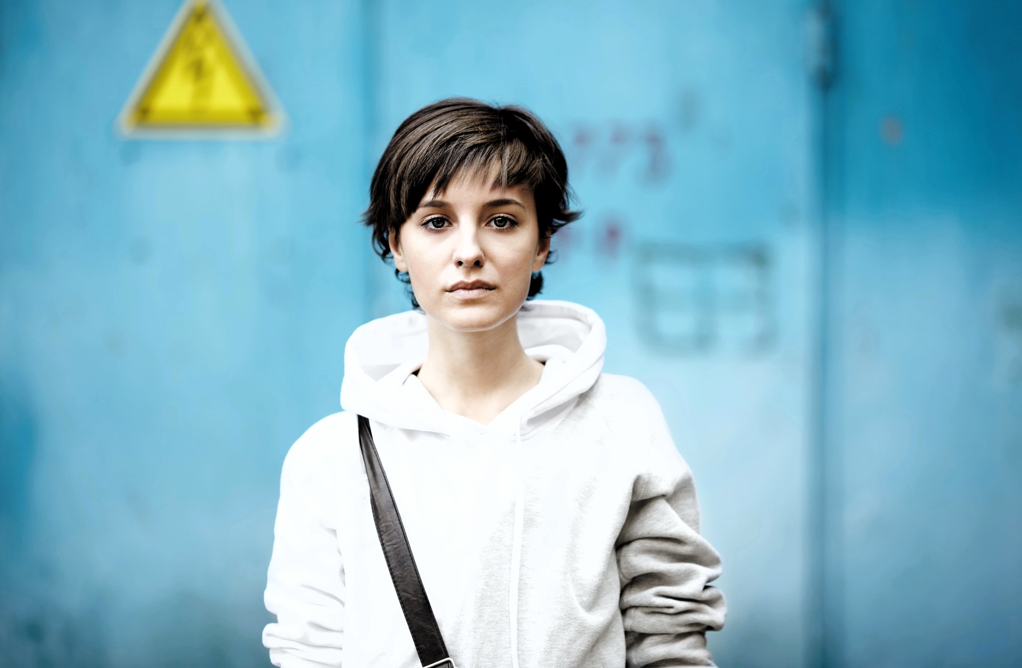 A person with short, dark hair wearing a white hoodie and a black strap across their shoulder stands in front of a blue wall with a yellow warning sign. The background is slightly out of focus.