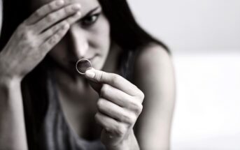 A woman with a pensive expression holds a ring in her hand. Her other hand rests on her forehead, partially obscuring her face. The image is in black and white, suggesting a somber or reflective mood.