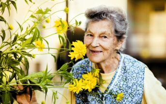 An elderly woman with gray hair smiles while holding a bouquet of bright yellow flowers next to her face. She wears a patterned blue and white vest over a yellow shirt. The background is softly blurred.