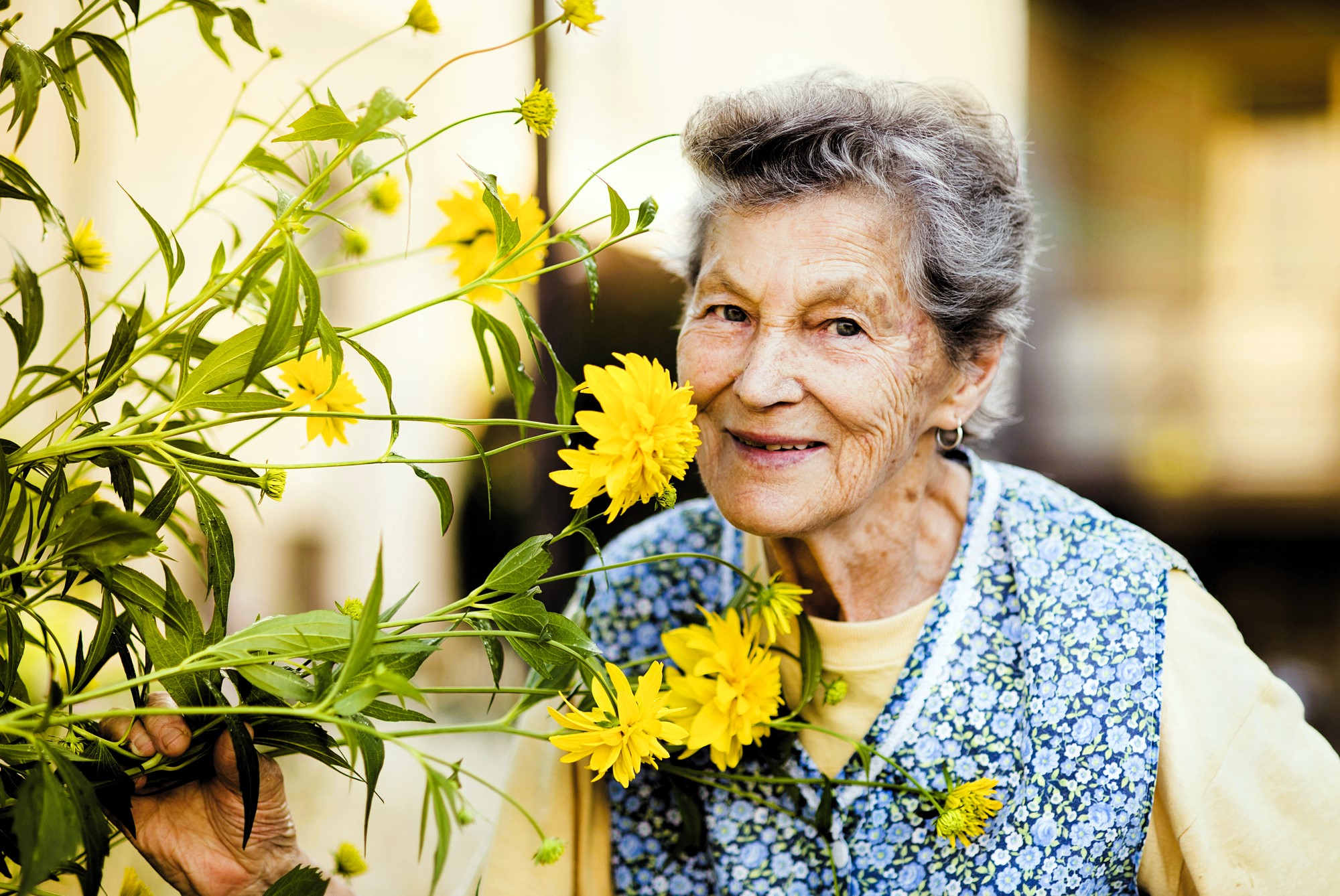 An elderly woman with gray hair smiles while holding a bouquet of bright yellow flowers next to her face. She wears a patterned blue and white vest over a yellow shirt. The background is softly blurred.