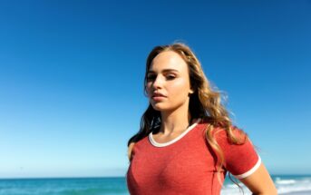 A woman with long hair stands on a beach, wearing a red shirt with white trim. The ocean and a clear blue sky are in the background.