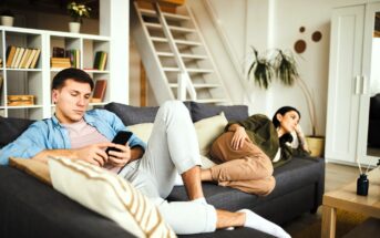 A man and a woman sit on a gray couch in a living room. The man is using a smartphone, wearing a blue shirt and light pants. The woman, in an olive jacket and brown pants, reclines nearby, looking away. Shelves and a staircase are in the background.