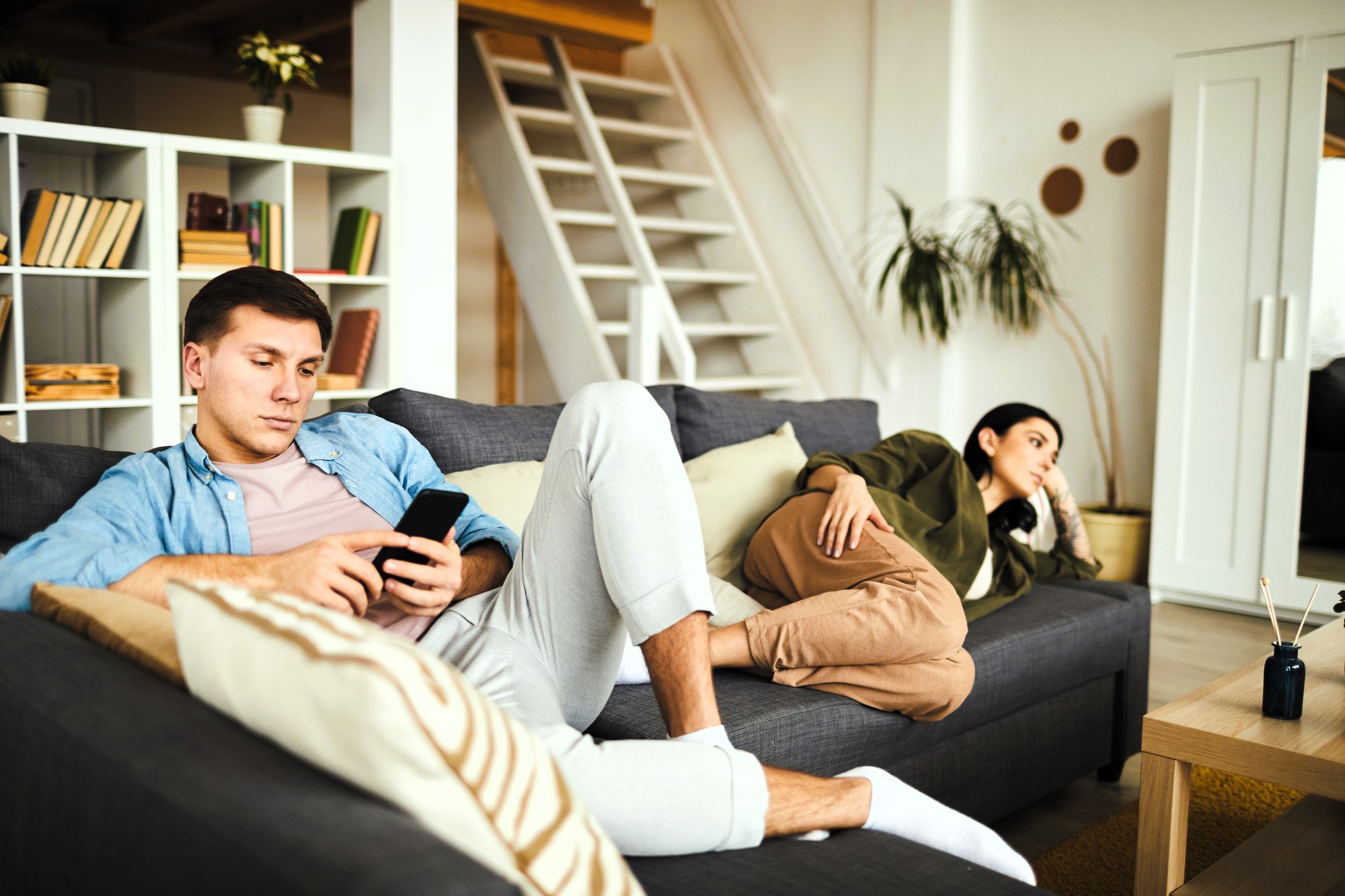 A man and a woman sit on a gray couch in a living room. The man is using a smartphone, wearing a blue shirt and light pants. The woman, in an olive jacket and brown pants, reclines nearby, looking away. Shelves and a staircase are in the background.