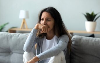 A woman is sitting on a gray couch with a thoughtful expression, resting her chin on her hand. She is wearing a white t-shirt and gray cardigan. In the background, there's a lamp and a potted plant on a wooden table.