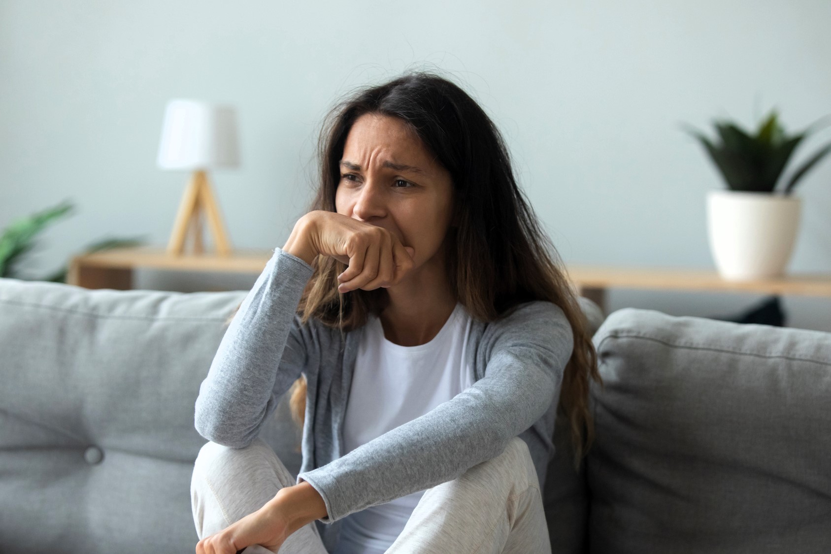 A woman is sitting on a gray couch with a thoughtful expression, resting her chin on her hand. She is wearing a white t-shirt and gray cardigan. In the background, there's a lamp and a potted plant on a wooden table.