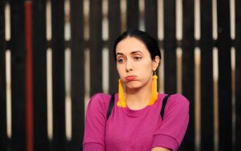 A person wearing a purple top and large yellow tassel earrings stands in front of a dark wooden fence, puffing their cheeks and looking to the side.