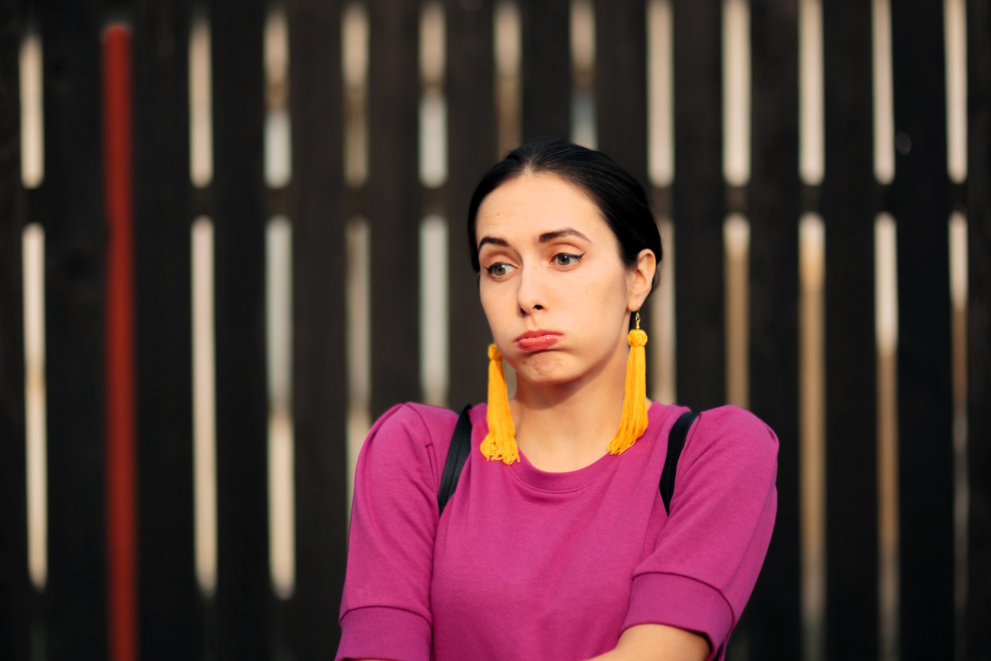 A person wearing a purple top and large yellow tassel earrings stands in front of a dark wooden fence, puffing their cheeks and looking to the side.