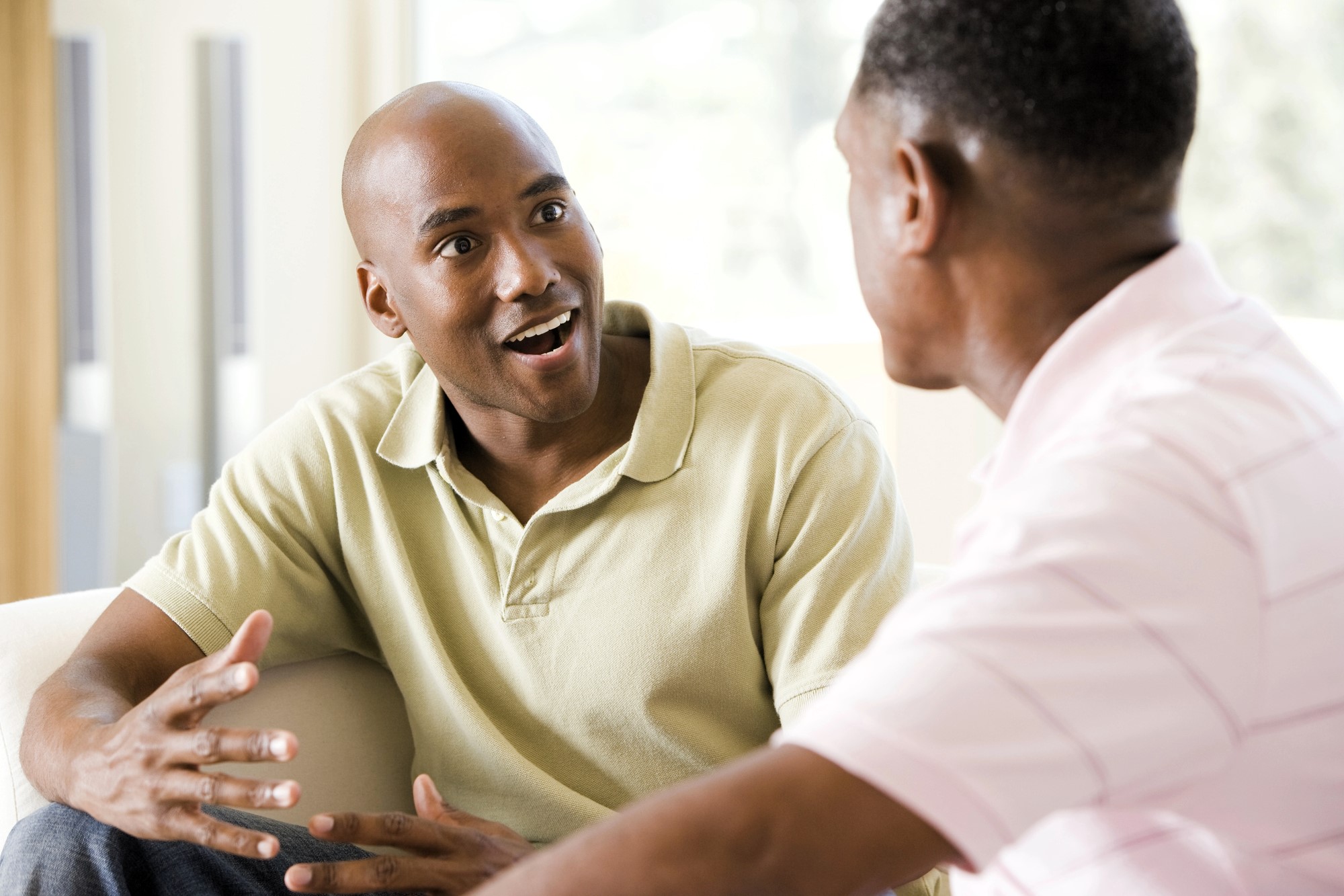 Two men are sitting indoors, engaged in a conversation. The man on the left, in a light green polo shirt, appears surprised or animated, gesturing with his hands. The man on the right is wearing a light pink shirt and facing away from the camera.