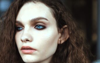 A close-up of a person with wavy brown hair and bright blue eyes, looking to the side. The lighting highlights smooth skin and subtle makeup, emphasizing the eyes and lips. The background is softly blurred.