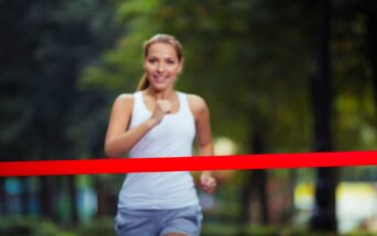 A woman in a white tank top and gray shorts runs toward the camera, smiling, as she crosses a red finish line in a park. The background is blurred with greenery.