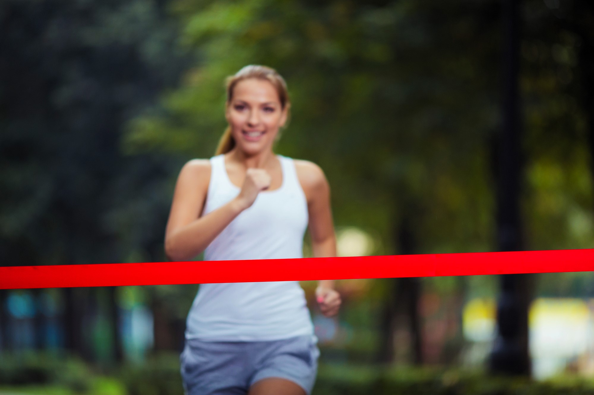 A woman in a white tank top and gray shorts runs toward the camera, smiling, as she crosses a red finish line in a park. The background is blurred with greenery.