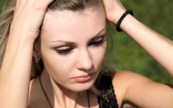 A woman stands outdoors with her hands on her head, eyes gently closed. Her hair is pulled back by her hands, and she wears a black top and a bracelet. The background is a blurred green, suggesting grass or foliage.