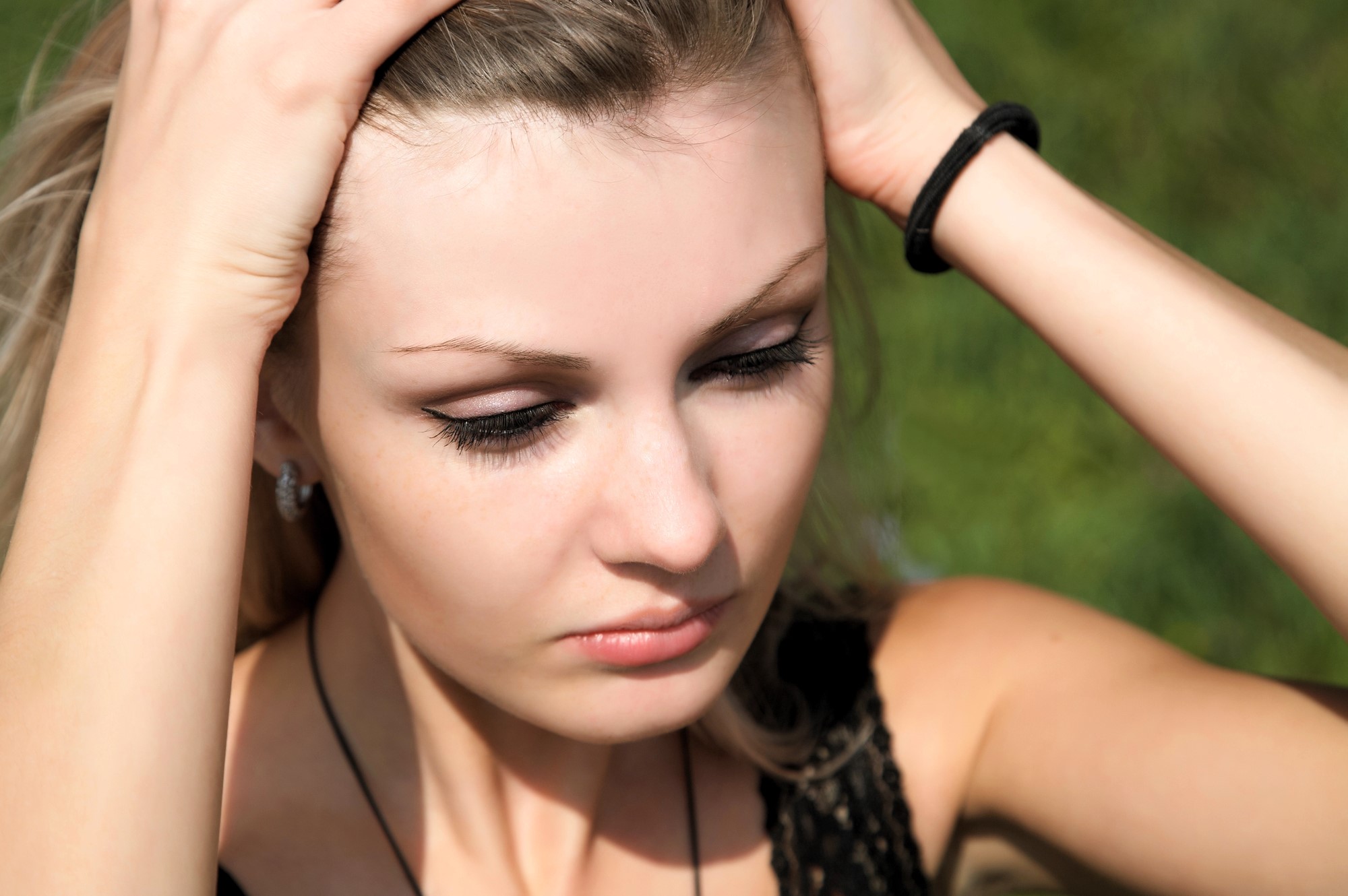 A woman stands outdoors with her hands on her head, eyes gently closed. Her hair is pulled back by her hands, and she wears a black top and a bracelet. The background is a blurred green, suggesting grass or foliage.