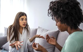 Two women sitting on a couch appear to be engaged in a heated conversation. The woman on the left is gesturing with her hands, looking surprised, while the woman on the right is also gesturing as they face each other.