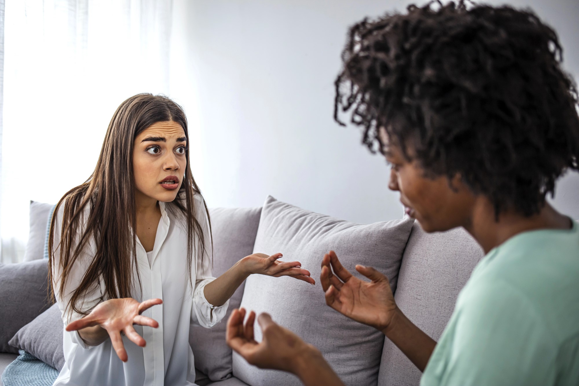 Two women sitting on a couch appear to be engaged in a heated conversation. The woman on the left is gesturing with her hands, looking surprised, while the woman on the right is also gesturing as they face each other.