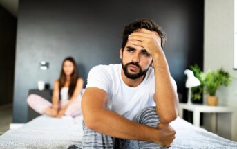 A man with a thoughtful expression sits on the edge of a bed, resting his head on his hand. He is wearing a white t-shirt and striped pants. In the background, a woman sits on the bed, slightly out of focus. A plant and lamp are beside her.