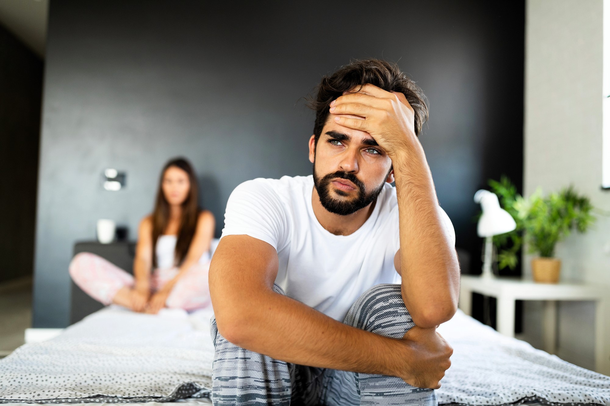 A man with a thoughtful expression sits on the edge of a bed, resting his head on his hand. He is wearing a white t-shirt and striped pants. In the background, a woman sits on the bed, slightly out of focus. A plant and lamp are beside her.