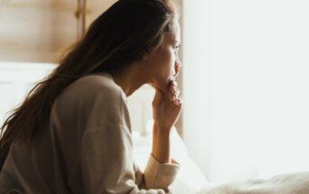 A woman with long hair sits indoors, looking thoughtfully out of a bright window. She rests her chin on her hand, wearing a cozy, light-colored sweater. The soft lighting creates a calm and reflective atmosphere.