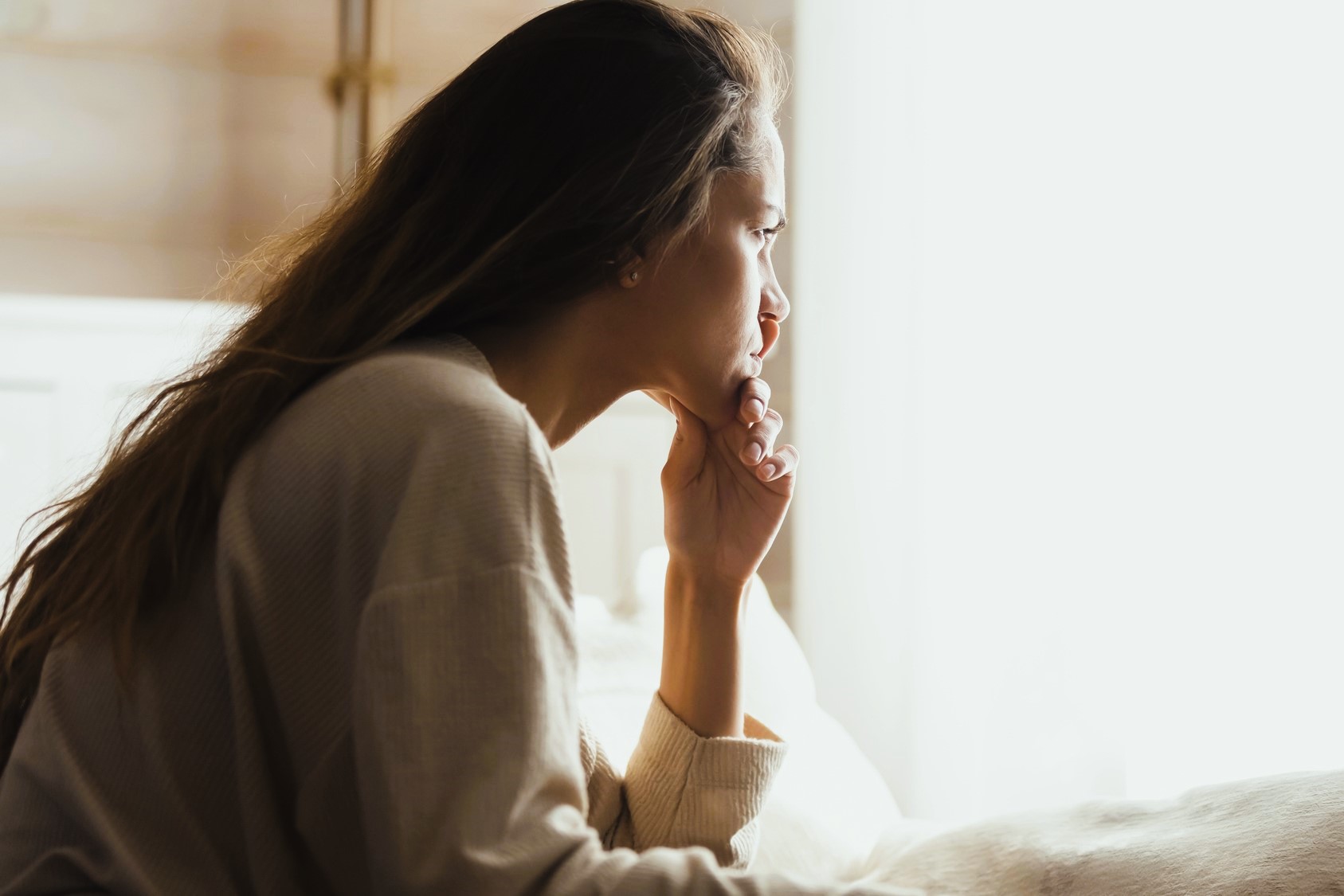 A woman with long hair sits indoors, looking thoughtfully out of a bright window. She rests her chin on her hand, wearing a cozy, light-colored sweater. The soft lighting creates a calm and reflective atmosphere.