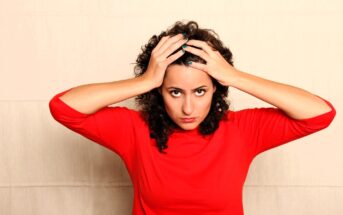 A woman with curly hair, wearing a red shirt, looks directly at the camera with a serious expression. Her hands are resting on her head against a plain background.