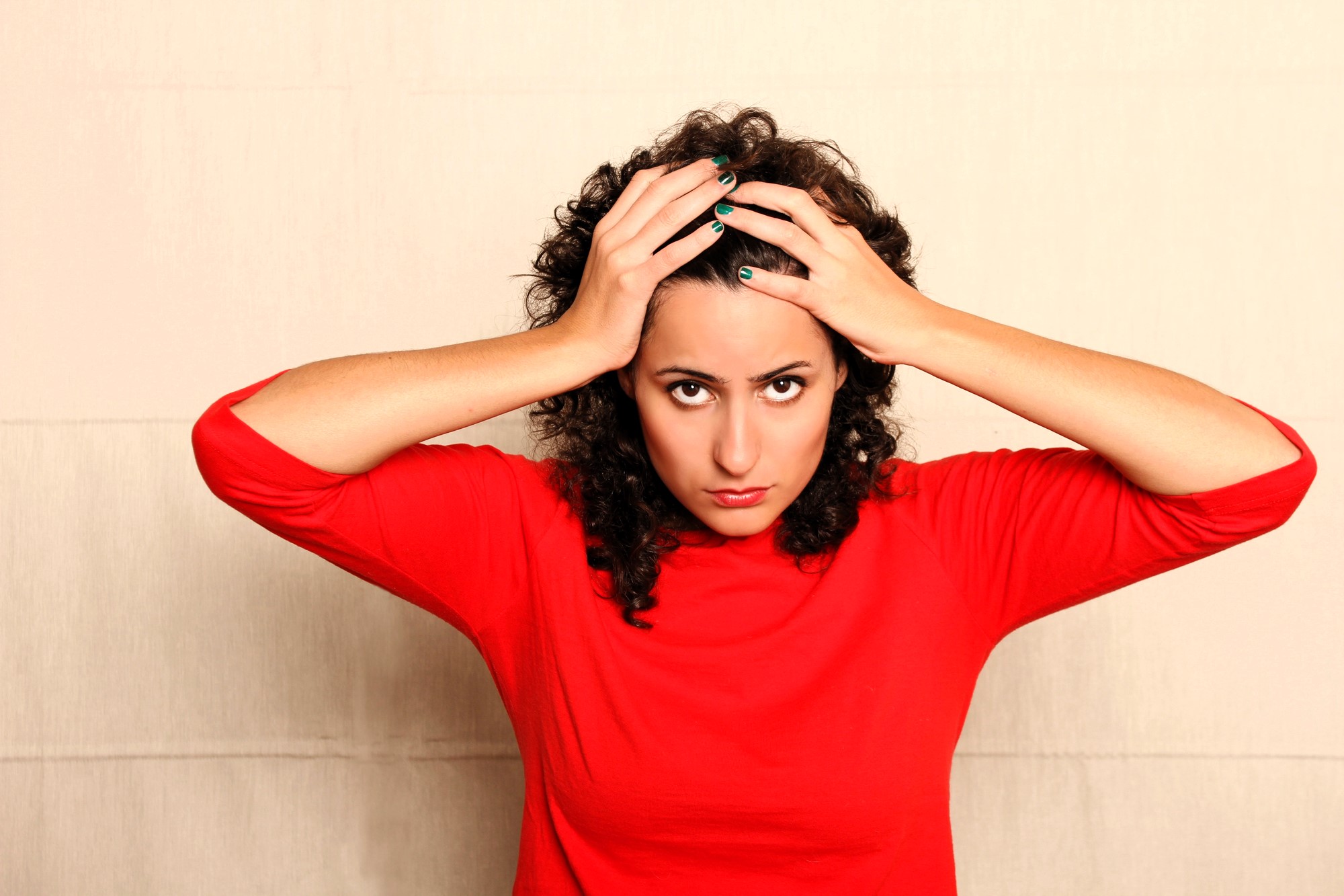 A woman with curly hair, wearing a red shirt, looks directly at the camera with a serious expression. Her hands are resting on her head against a plain background.