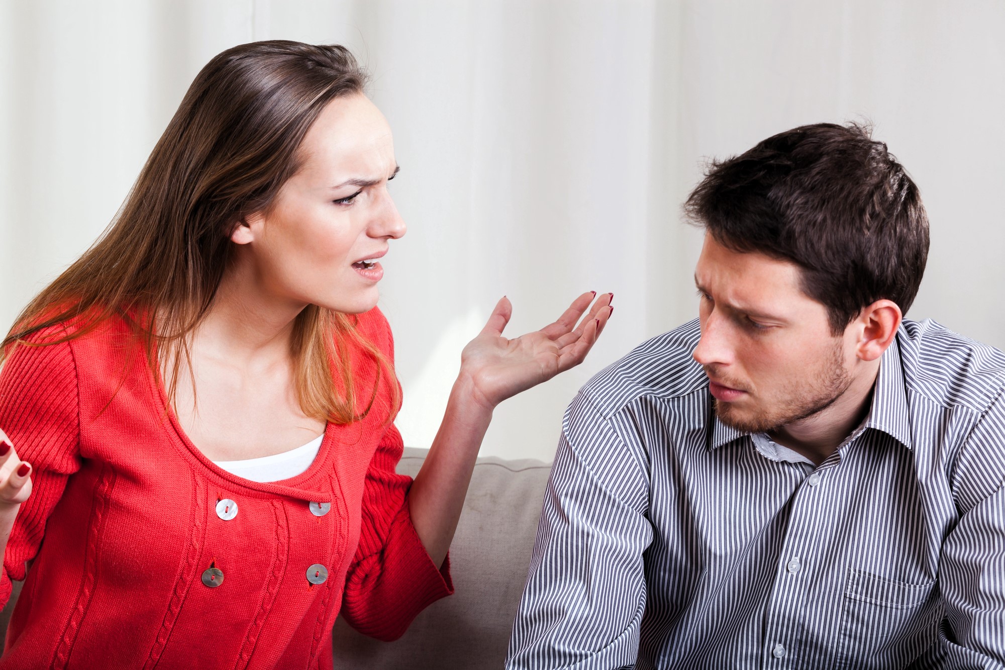 A woman wearing a red sweater is gesturing and looking upset while talking to a man in a striped shirt. The man is sitting beside her, looking down with a concerned expression. They are indoors against a light-colored background.