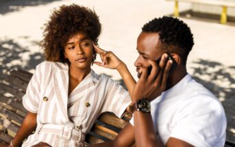 A woman and a man sitting on a bench outdoors. The woman, wearing a striped dress, rests her head on her hand, looking at the man. The man, in a white shirt, is holding a phone to his ear. They are surrounded by sunlight and shadows.