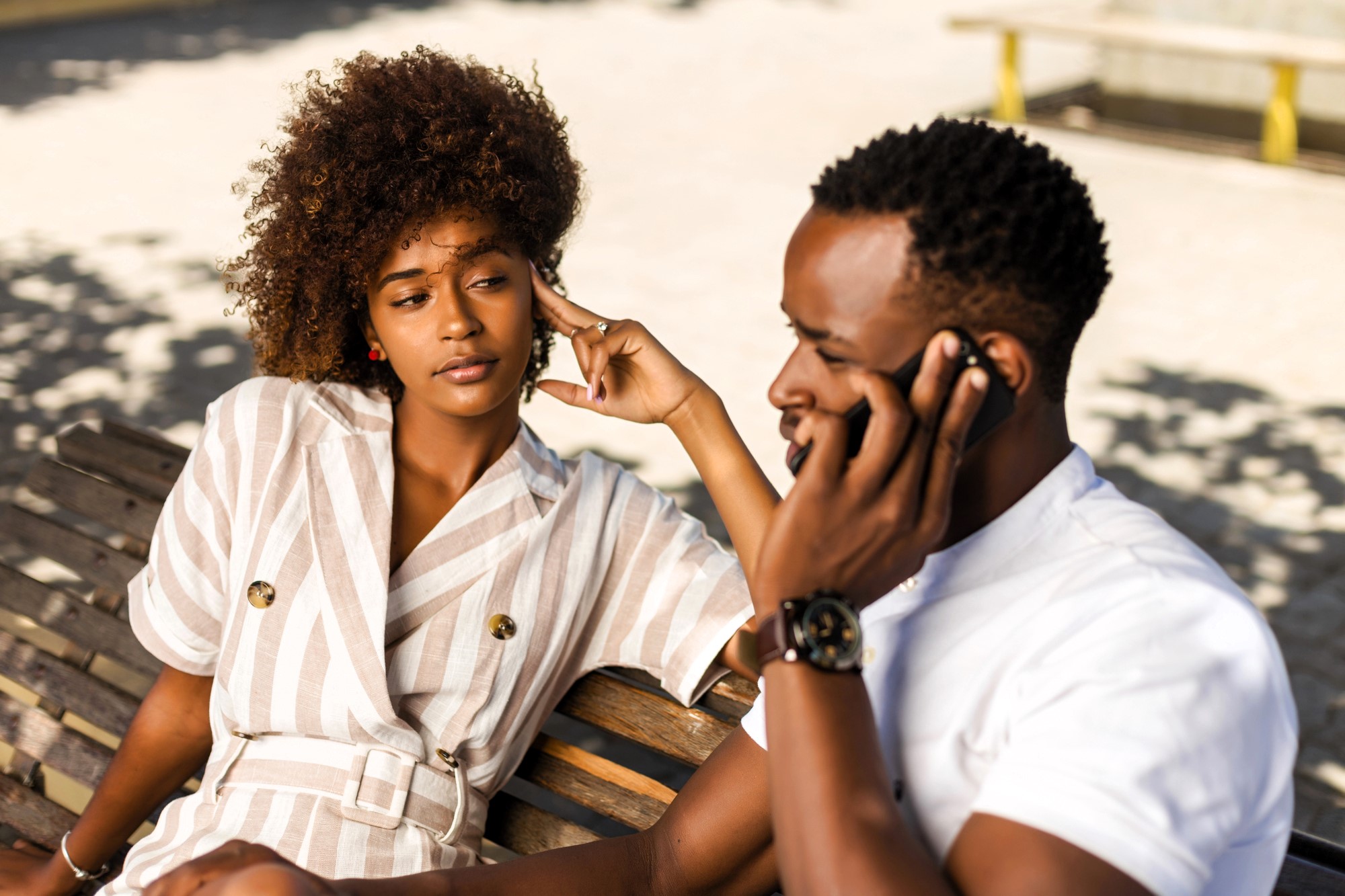 A woman and a man sitting on a bench outdoors. The woman, wearing a striped dress, rests her head on her hand, looking at the man. The man, in a white shirt, is holding a phone to his ear. They are surrounded by sunlight and shadows.