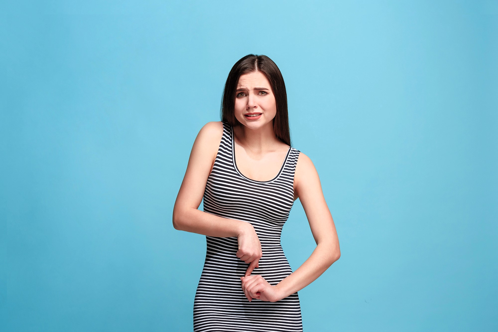 A woman in a black and white striped dress stands against a blue background. She has long, straight hair and is looking to the side with a slightly confused or unsure expression, with her arms bent at her sides.