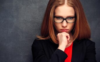 A woman with long auburn hair and glasses looks down pensively, resting her chin on her hand. She is wearing a black blazer over a red top, with a dark, textured background behind her.