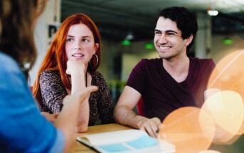 Three people are engaged in a discussion at a table indoors. Two people are facing each other; one with red hair listening attentively and another with dark hair smiling. A person in the foreground gestures while speaking. The table has an open book.