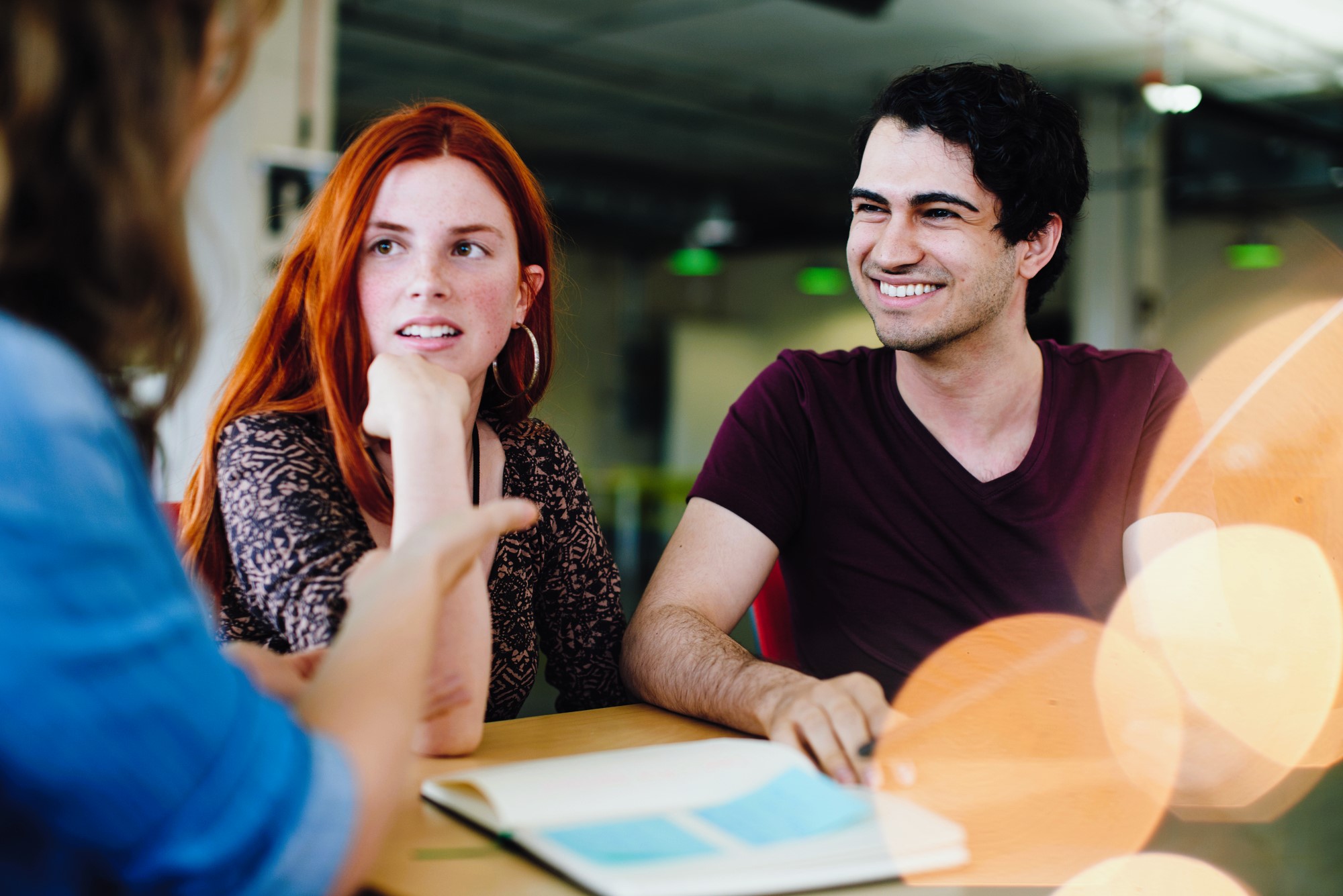 Three people are engaged in a discussion at a table indoors. Two people are facing each other; one with red hair listening attentively and another with dark hair smiling. A person in the foreground gestures while speaking. The table has an open book.