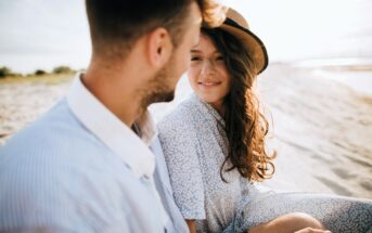 A couple sitting on a sandy beach. The woman with long brown hair is wearing a light patterned outfit and a hat, smiling at the man beside her. The man is wearing a blue and white striped shirt, and they're both enjoying a sunny day.