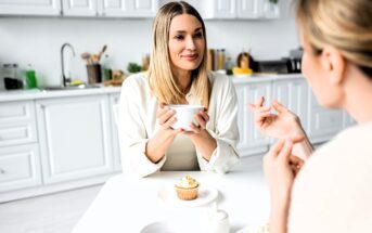 Two women are sitting at a white kitchen table having a conversation. One woman holds a mug, and there are cupcakes and tea on the table. The kitchen in the background is bright and modern.