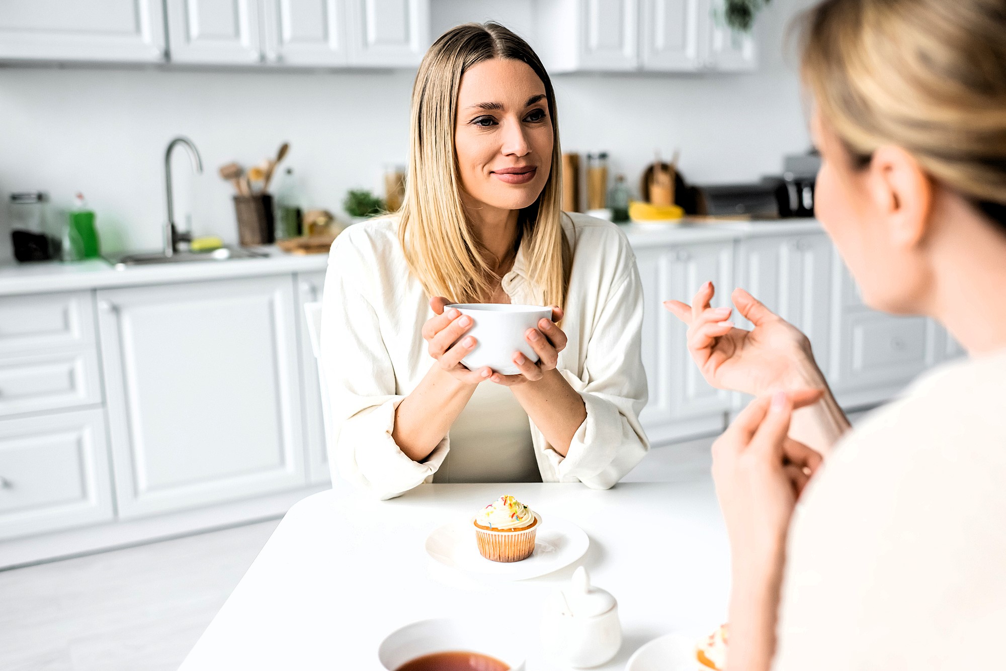 Two women are sitting at a white kitchen table having a conversation. One woman holds a mug, and there are cupcakes and tea on the table. The kitchen in the background is bright and modern.