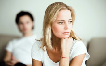 A woman with blond hair and a white shirt looks thoughtfully into the distance, resting her chin on her hand. In the blurred background, a person with dark hair and a white shirt is seated. The mood appears contemplative.