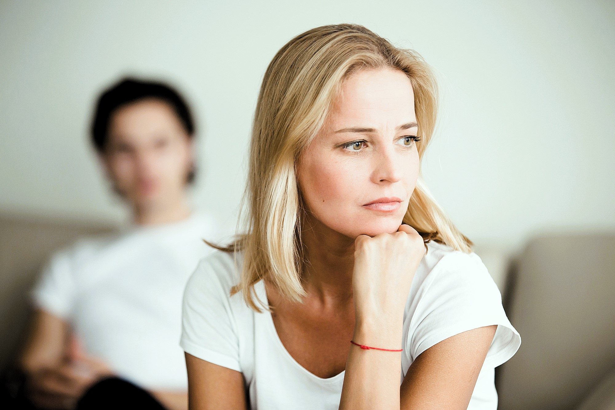 A woman with blond hair and a white shirt looks thoughtfully into the distance, resting her chin on her hand. In the blurred background, a person with dark hair and a white shirt is seated. The mood appears contemplative.