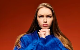 A woman with long brown hair, wearing a blue sweater, clasps her hands under her chin, looking serious against a red background.