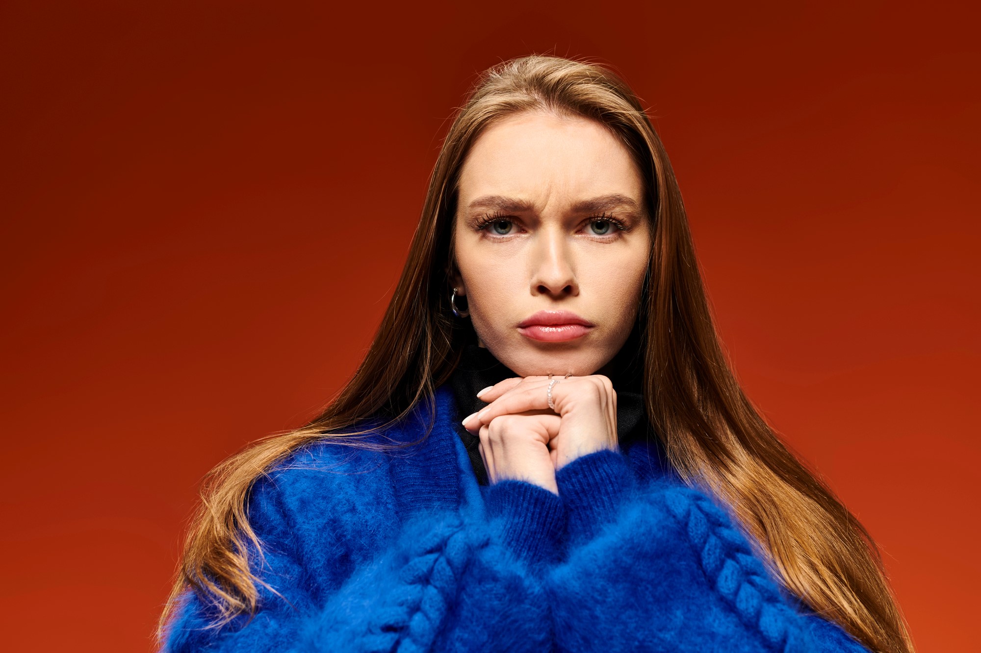 A woman with long brown hair, wearing a blue sweater, clasps her hands under her chin, looking serious against a red background.