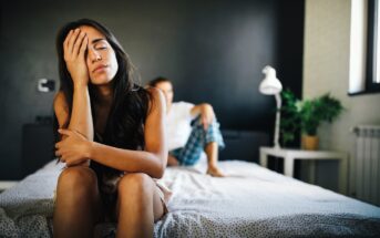 A woman sits on the edge of a bed, her hand on her forehead, looking distressed. In the background, a person is sitting on the bed, partially out of focus. The room is dimly lit, with a plant and lamp visible.