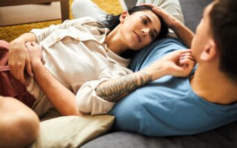 A couple relaxes on a couch, with the woman lying on her back, looking up at the man, who is sitting and has a tattooed arm. They share an intimate and peaceful moment, surrounded by soft furnishings in a cozy setting.